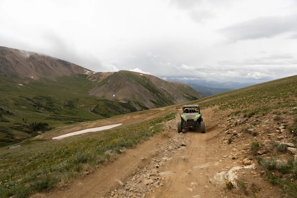 Green Kawasaki KRX side-by-side driving an alpine dirt road toward distant peaks