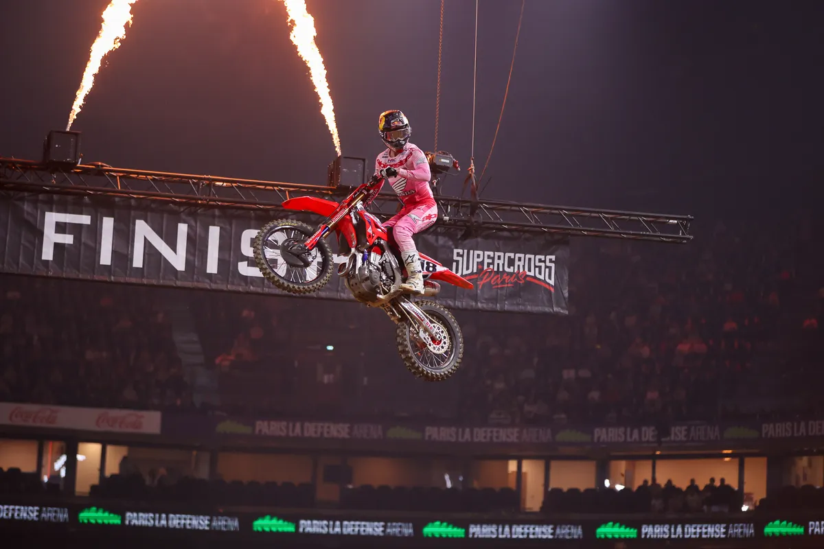 Supercross rider jumping a red Honda past the finish-line banner as flames shoot from the gantry above