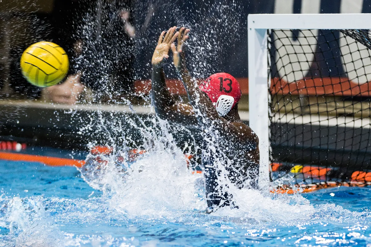 Goalkeeper in a red cap lunging up through a wall of spray as the ball flies toward the net