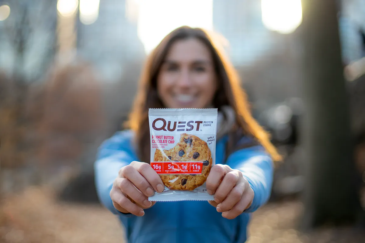 Olivia holding a Quest protein cookie toward the camera in a park at sunset