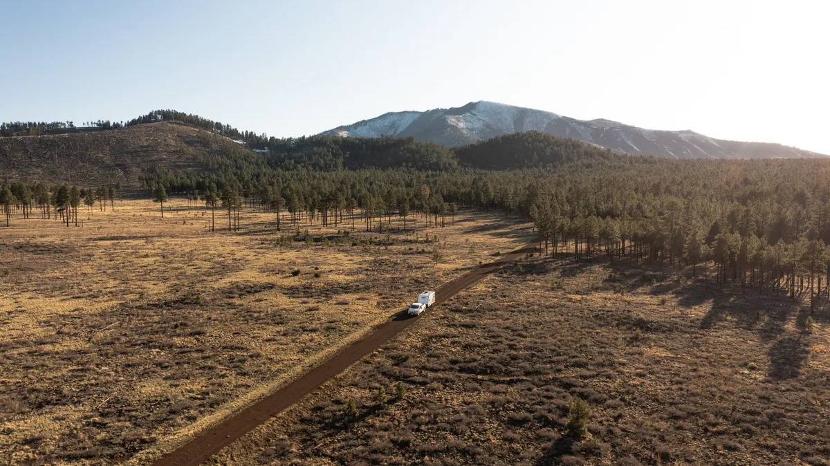 Aerial of a pickup towing a travel trailer along a dirt road through a valley below a snow-dusted mountain