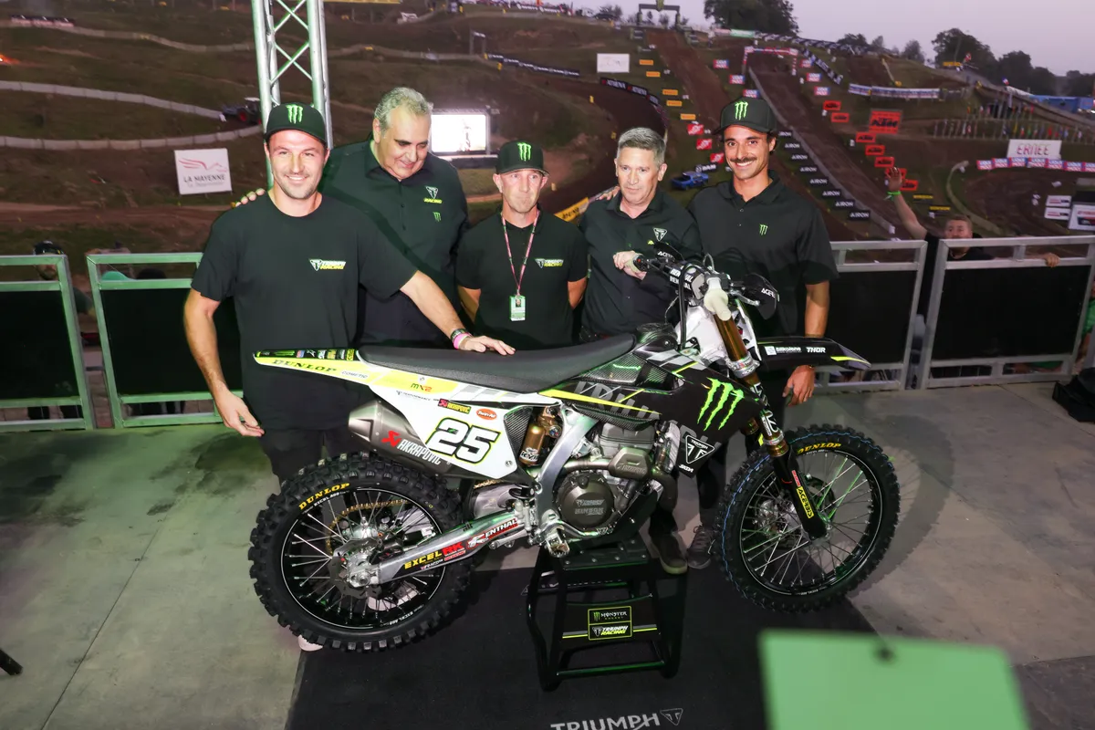 Team riders and staff gathered around the new Triumph motocross bike on stage at dusk, the track behind
