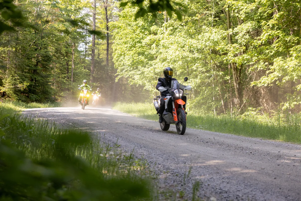 Adventure rider on a KTM kicking up dust along a sunlit gravel road through the forest, another rider following behind