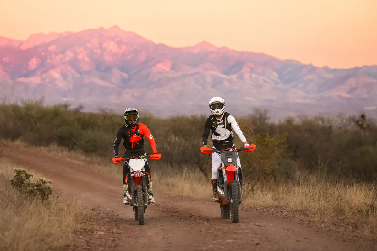 Two dirt bike riders on a dirt road at dusk with pink light on the mountains behind