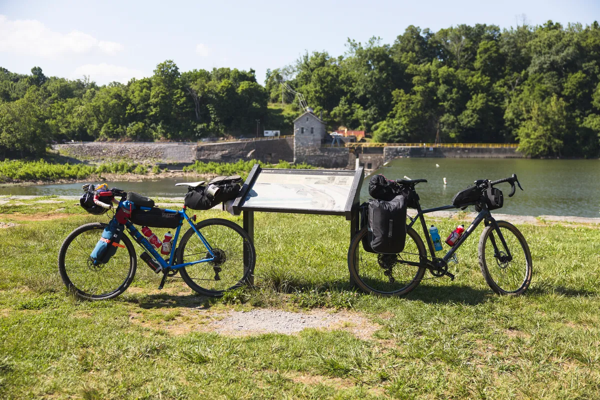 Two loaded bikes leaning against an interpretive sign by the river, with a dam in the background