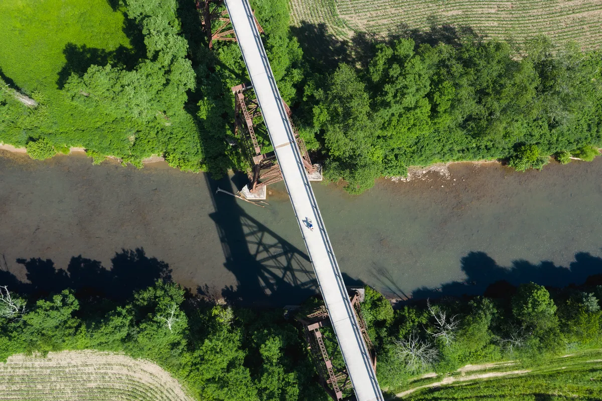 Top-down drone view of a trail bridge over a river with a tiny rider on the deck