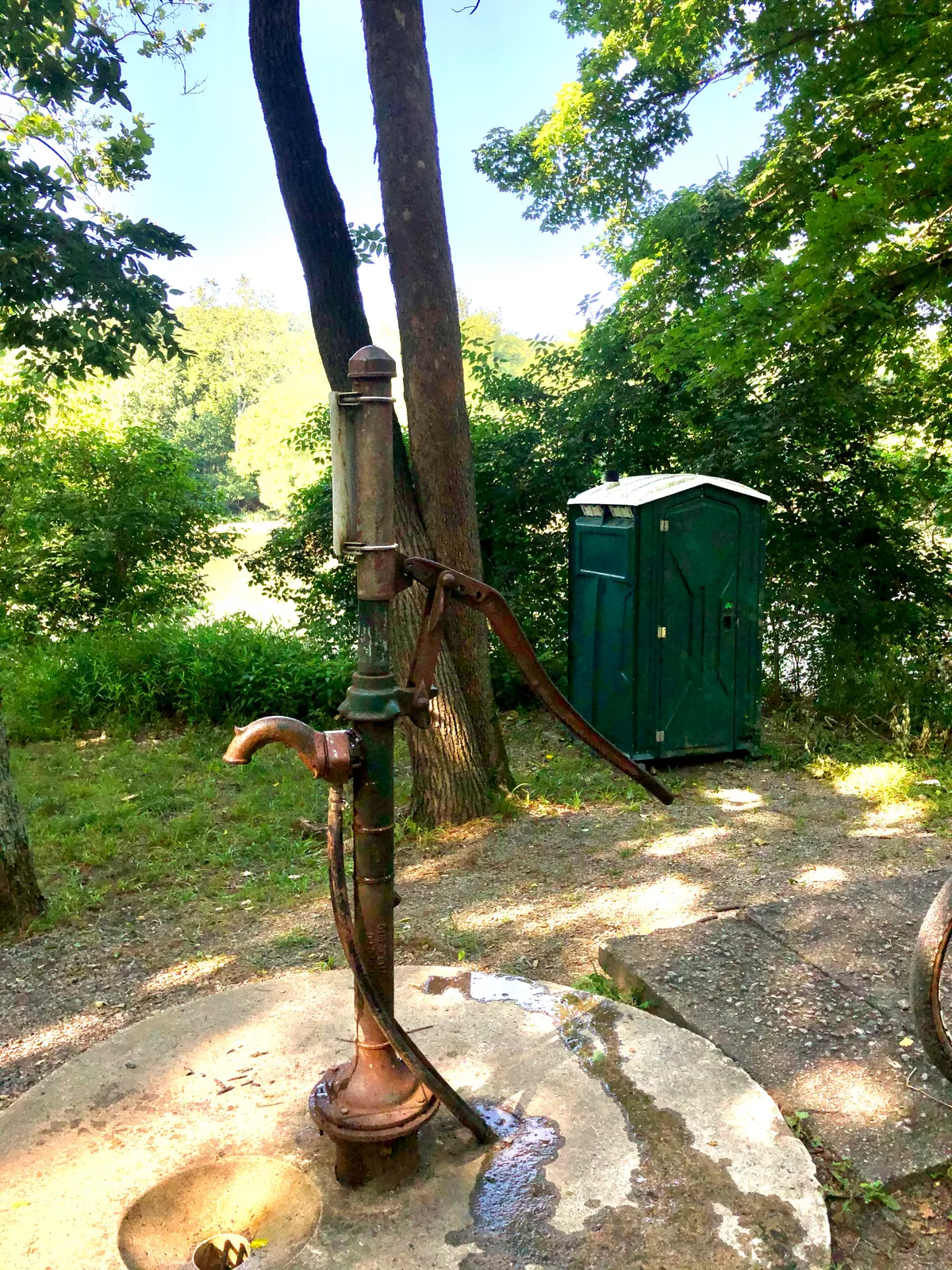 Hand water pump at a C&O campsite with a green porta-potty behind