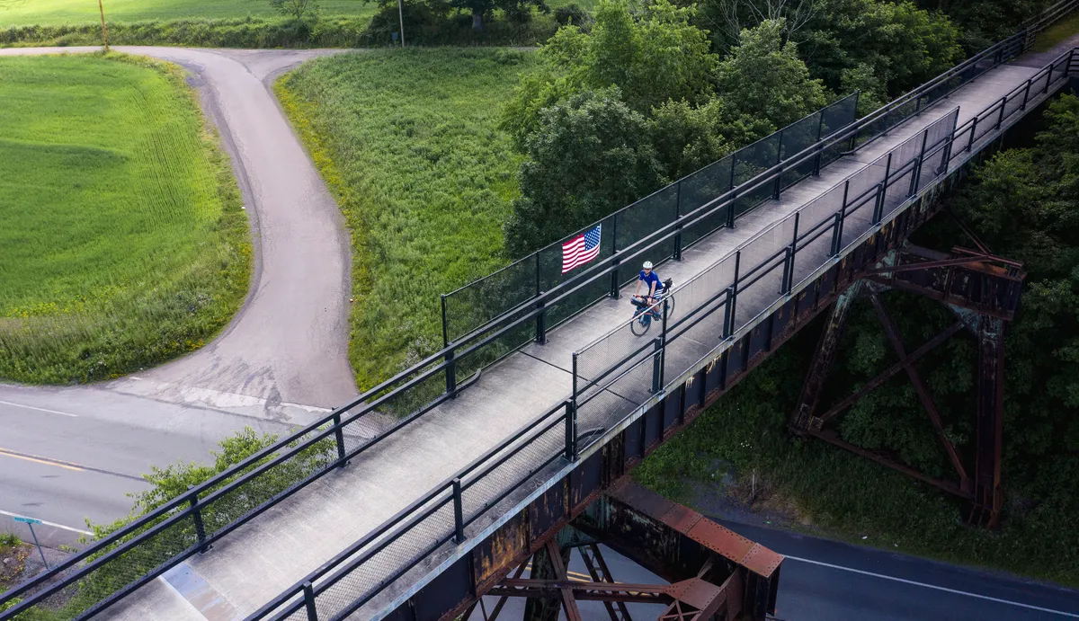Drone view of a rider crossing a trail bridge with an American flag on the railing, road curving below
