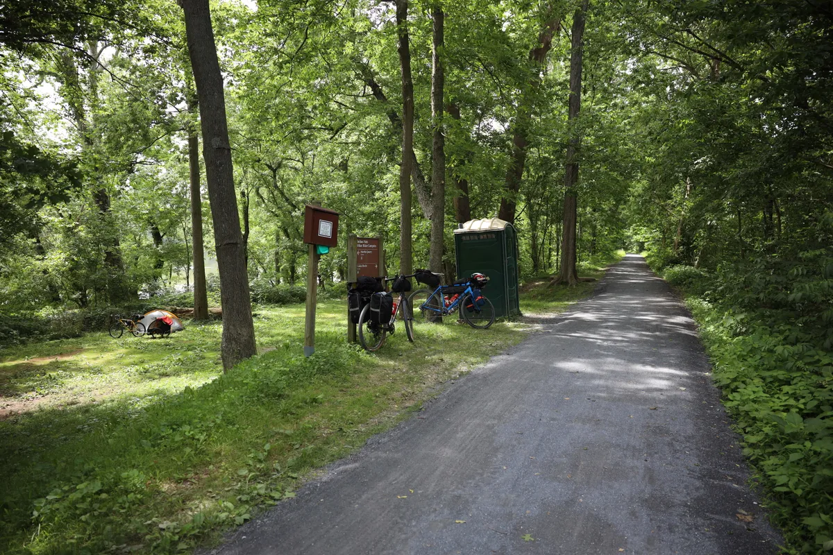 Hiker/biker campsite right off the gravel towpath with signposts, bikes, a porta-potty and a tent in the trees