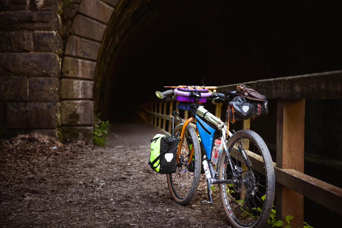 Loaded gravel bike with panniers and a bar bag leaning on the wooden railing at the mouth of the Paw Paw tunnel