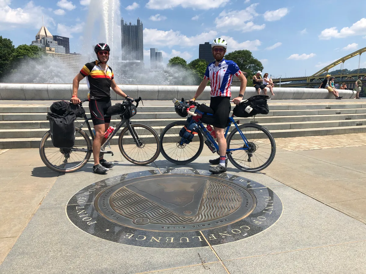 Alex and Dan with their loaded bikes at the Point State Park fountain, downtown towers and a yellow bridge behind