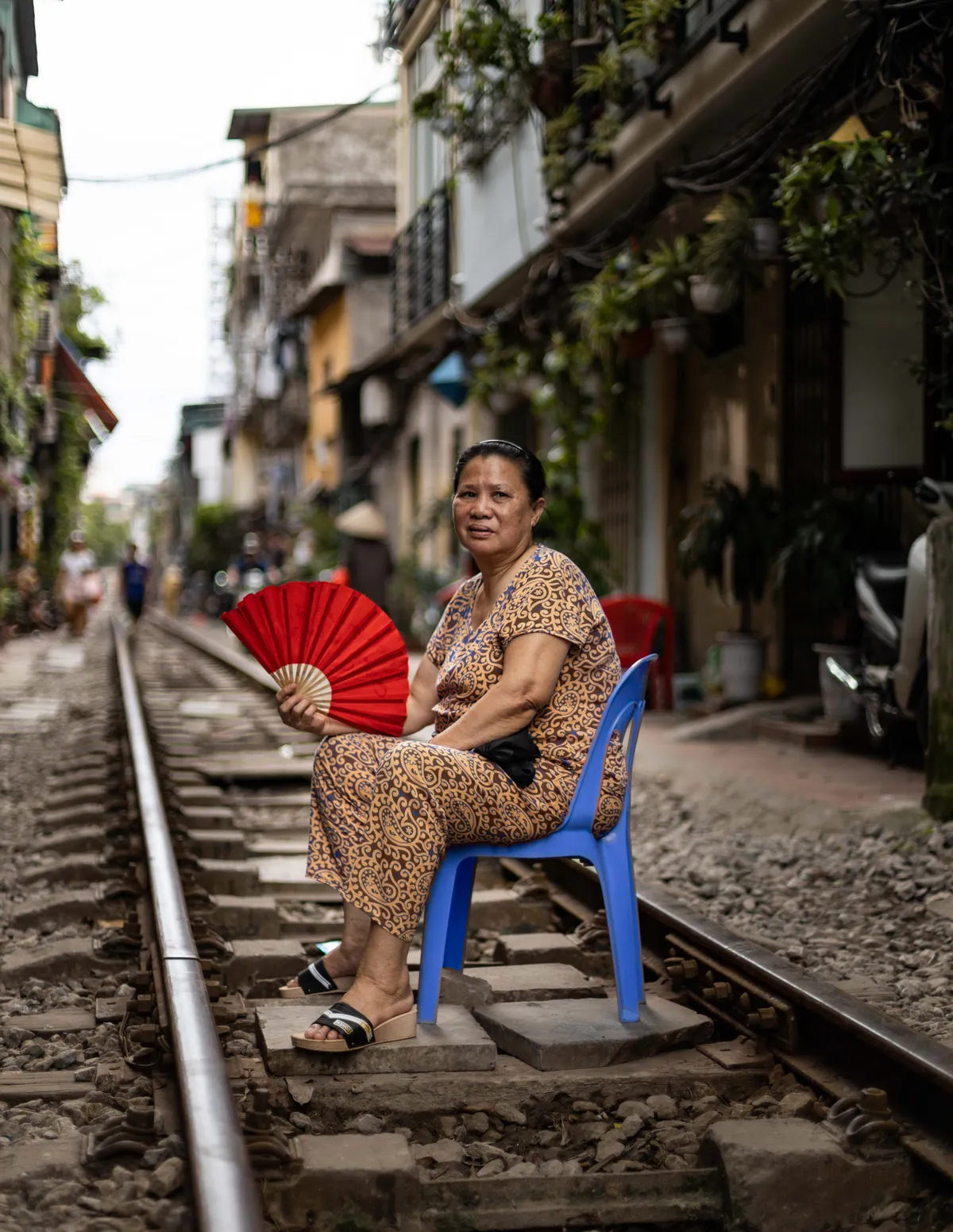 Woman with a red fan sitting on a blue chair beside the tracks on Train Street
