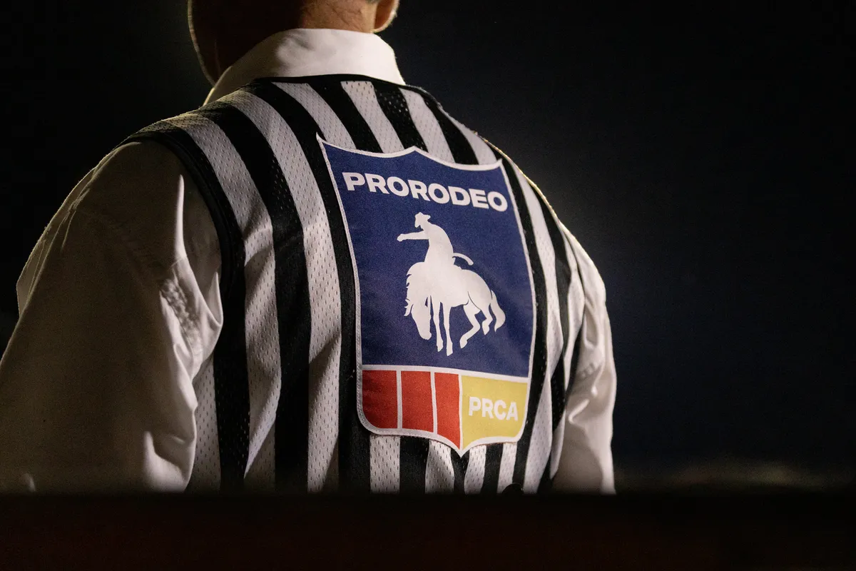 Rodeo judge in a striped shirt with a PRORODEO patch, seen from behind against a dark background