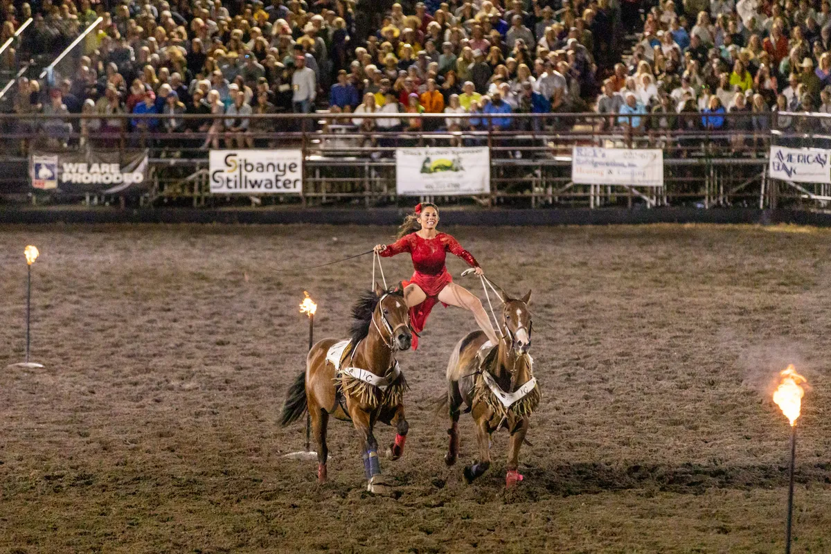 Trick rider in red standing on two horses between flaming torches at night