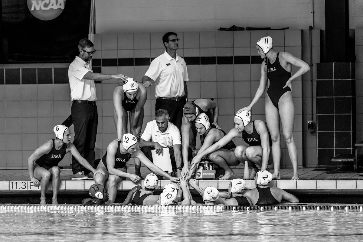 Black and white photo of USA players and coaches stacking hands at the pool edge