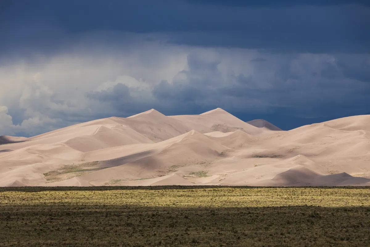 Pale sand dunes under a dark storm sky, lit by a band of sun across the grassland in front