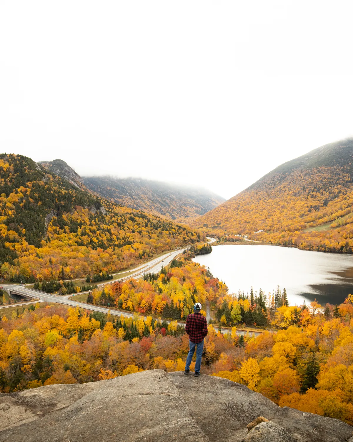 Hiker in a plaid shirt standing on a rock ledge above a lake and highway winding through a valley of autumn foliage