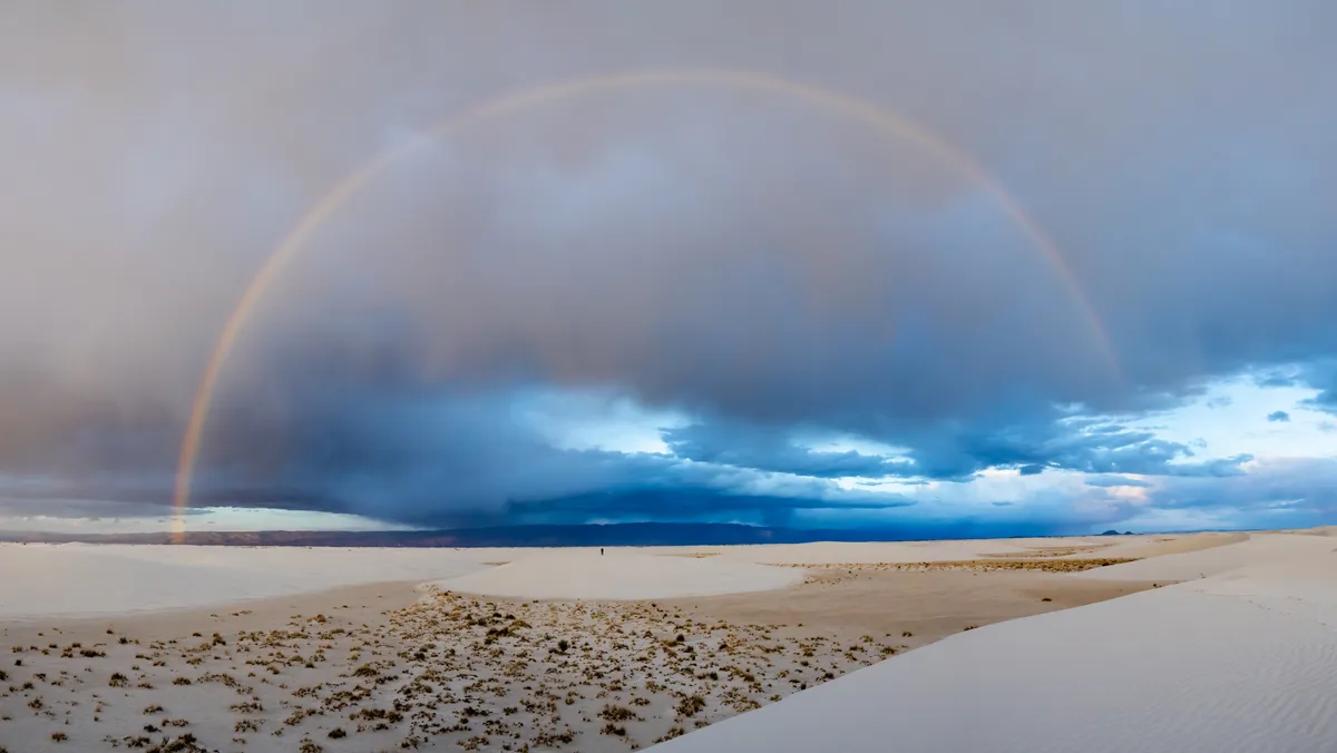 A full rainbow arcs over a storm front above white gypsum dunes and desert scrub