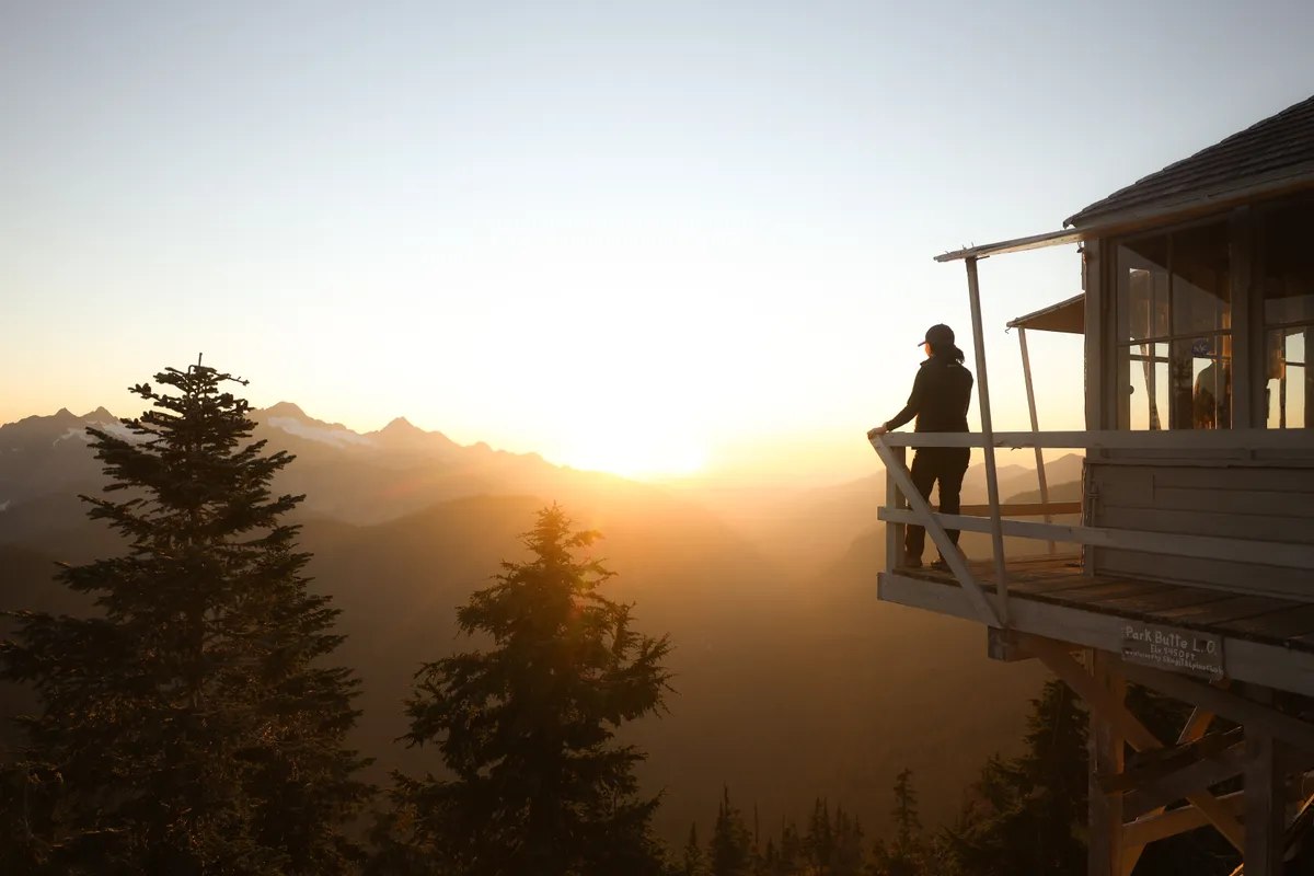 Person standing on the deck of a mountain fire lookout as the sun sets over the peaks