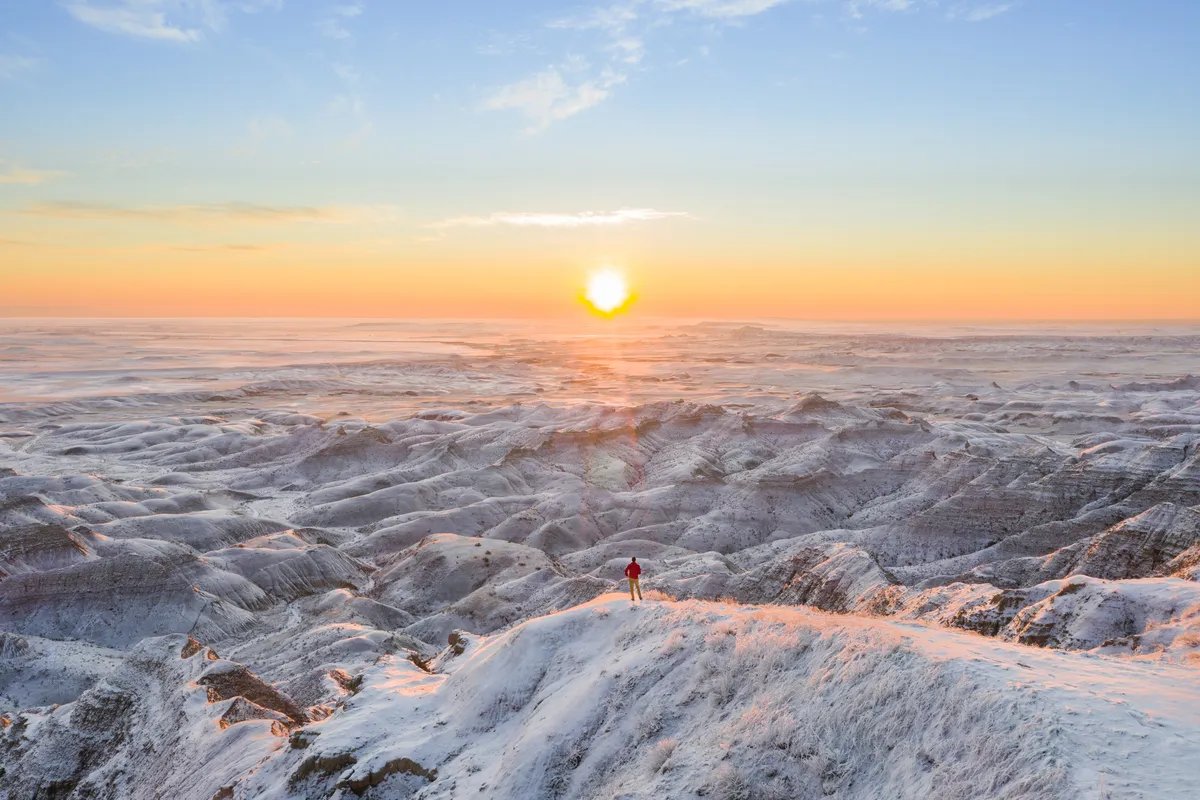 Lone hiker on a snowy ridge as the sun rises over a vast frosted badlands landscape