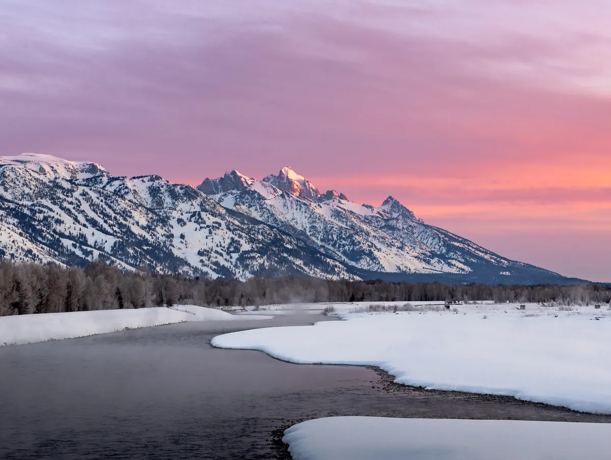 Pink alpenglow over the snowy Teton Range above a partly frozen river